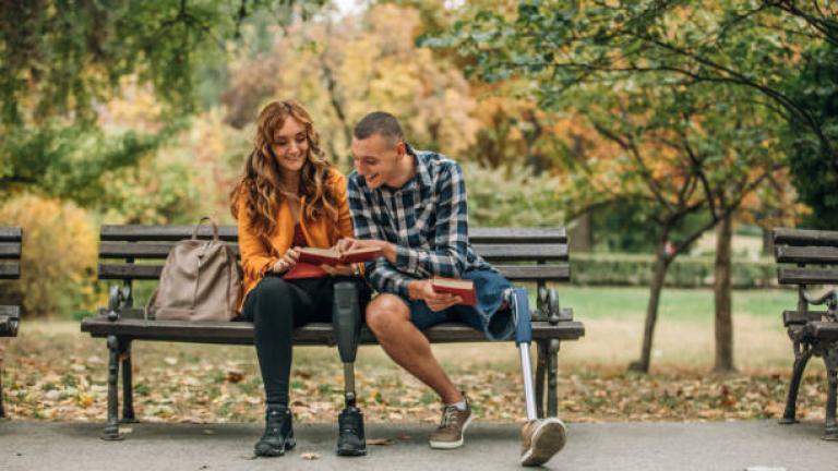 Two people sit on a bench. Girl and guy in early 30s. They look romantic on a bench with a park scene behind them. They both are missing legs and show prosthetics.