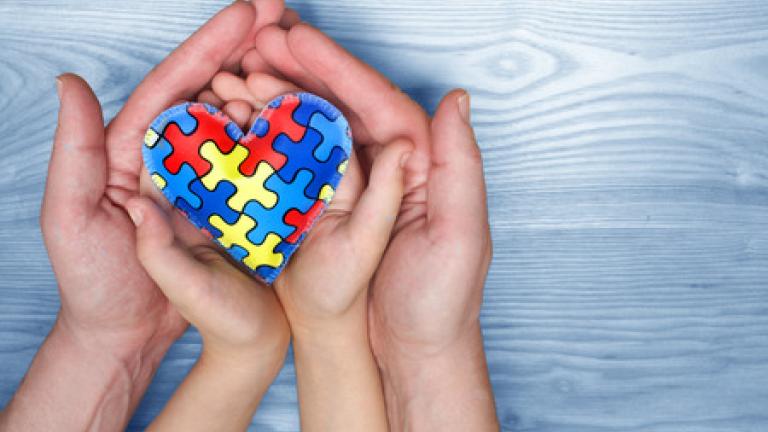 Adult hands cupping child hands. The child's hands are holding a heart made out of puzzle pieces