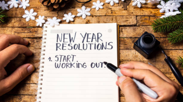 A pair of hands, one holding a pen over a pad, that reads &quot;New Year Resolutions, start working out&quot;